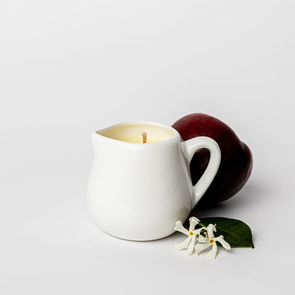 White mug with a lit candle inside, next to a red fruit and white flowers on a light gray background