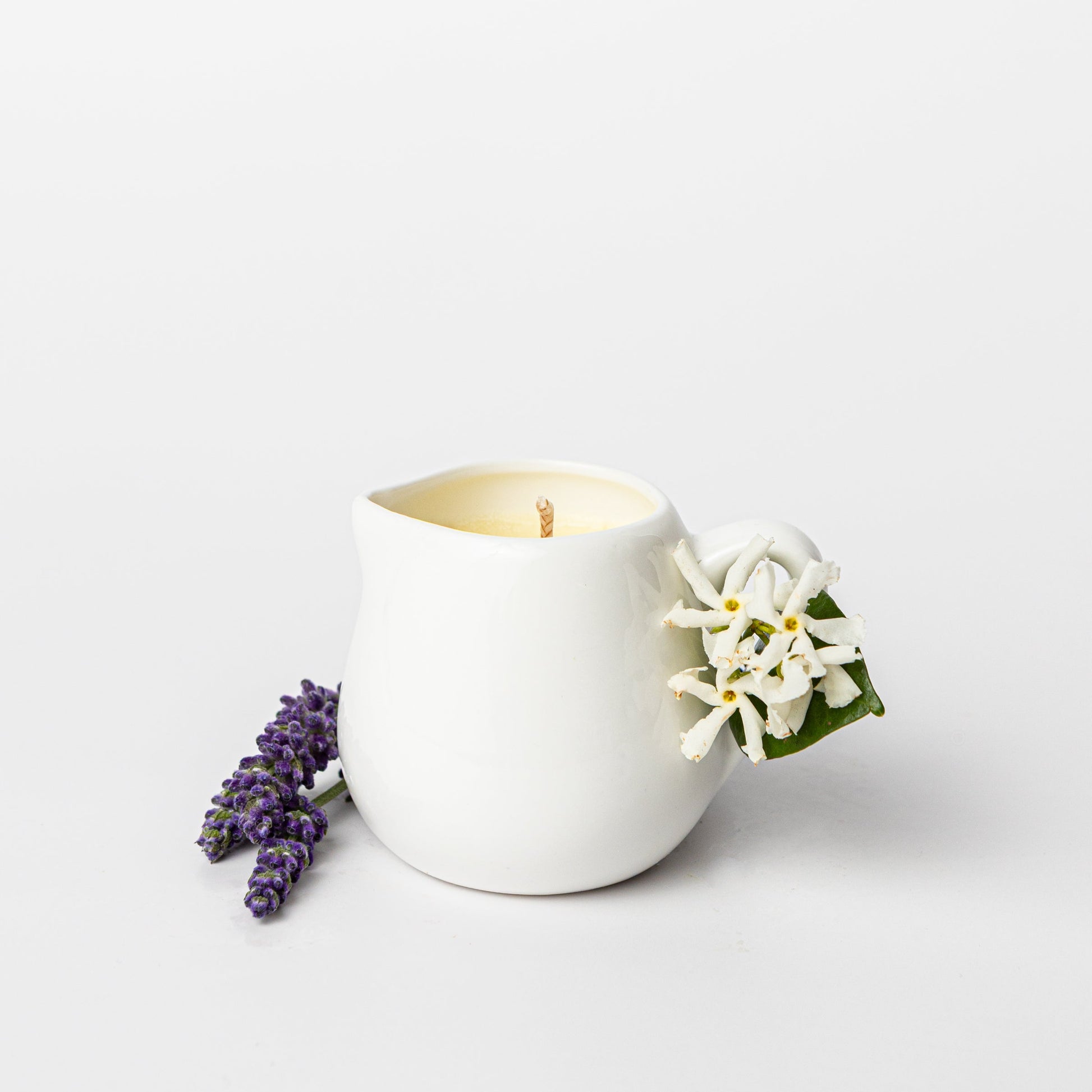 White ceramic candle holder with a lit candle, surrounded by lavender and white flowers on a light background